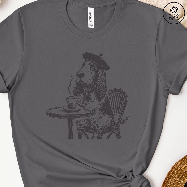 K&ouml;nnte beinhalten: Eine Schwarzwei&szlig;illustration eines Basset Hounds, der einen Baskenm&uuml;tze tr&auml;gt und an einem Tisch mit einer Tasse Kaffee sitzt. Der Hund sitzt auf einem Stuhl mit einer Korbgeflecht-R&uuml;ckenlehne.