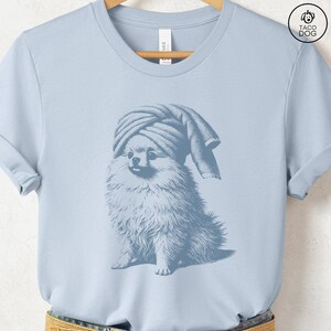 May include: Light blue t-shirt featuring a Pomeranian dog wearing a towel on its head. The dog illustration is in a vintage style, with detailed fur and a relaxed pose. The shirt has a classic crew neck and short sleeves.
