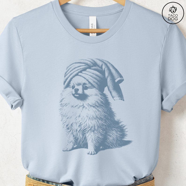 May include: Light blue t-shirt featuring a Pomeranian dog wearing a towel on its head. The dog illustration is in a vintage style, with detailed fur and a relaxed pose. The shirt has a classic crew neck and short sleeves.