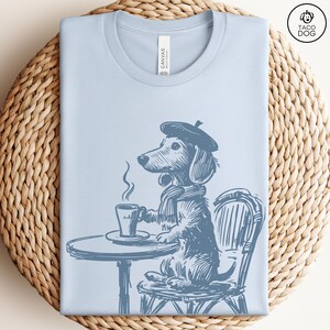 Może przedstawiać: Jasno niebieski t-shirt z grafiką przedstawiającą jamnika w berecie i szaliku, siedzącego przy stole z filiżanką kawy. Na koszulce widoczny jest napis "Canvas".
