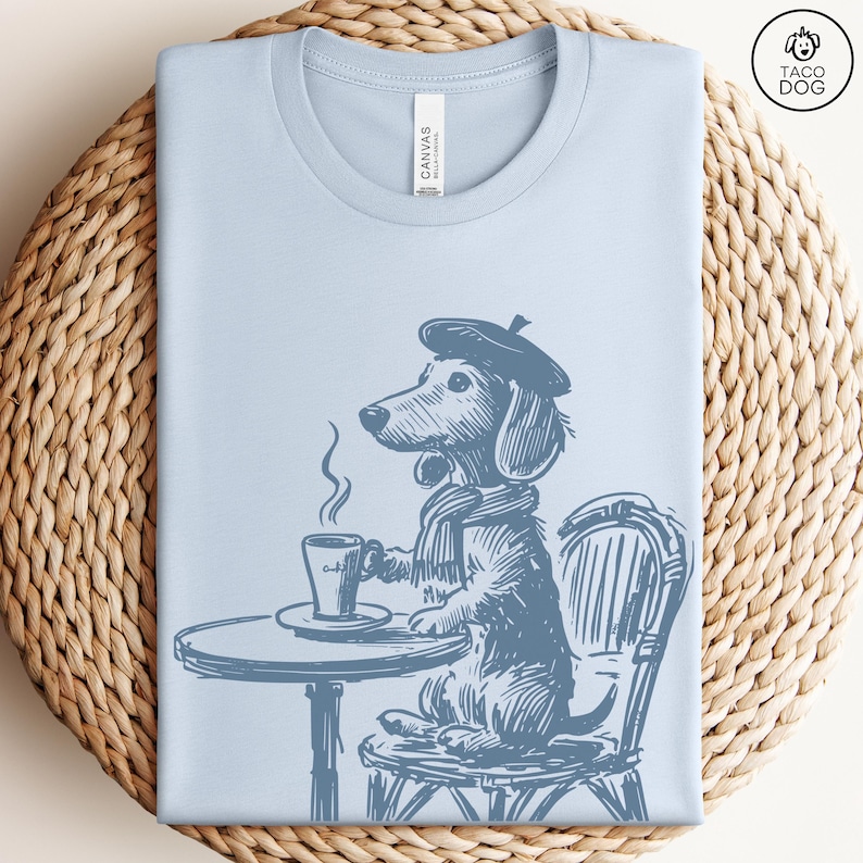 Może przedstawiać: Jasno niebieski t-shirt z grafiką przedstawiającą jamnika w berecie i szaliku, siedzącego przy stole z filiżanką kawy. Na koszulce widoczny jest napis "Canvas".