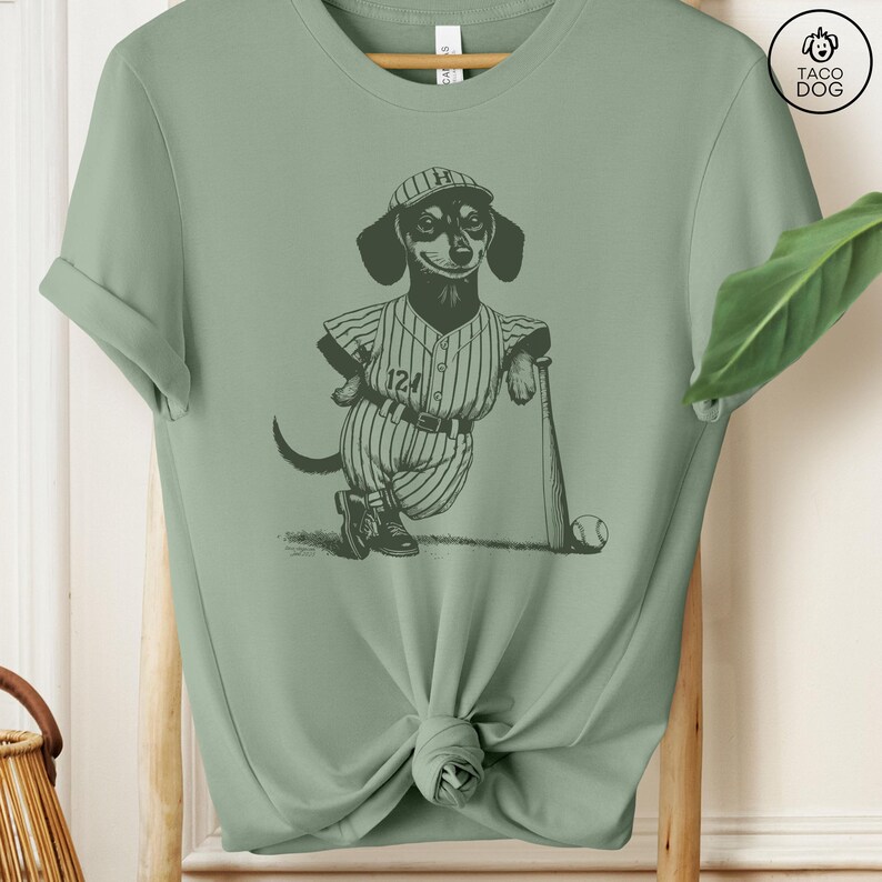May include: A sage green t-shirt with a black and white illustration of a dachshund wearing a baseball uniform and holding a bat. The dachshund is wearing a baseball cap with the letter "H" on it. The text "124" is on the uniform.
