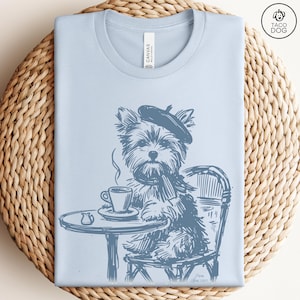 May include: Light blue t-shirt featuring a sketch of a Yorkshire Terrier wearing a beret and scarf, seated at a cafe table with a cup of coffee. The design is in shades of blue and includes the text "Taco Dog".