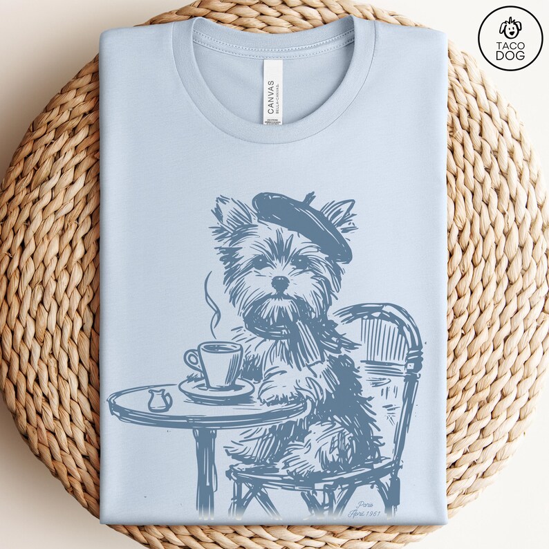 May include: Light blue t-shirt featuring a sketch of a Yorkshire Terrier wearing a beret and scarf, seated at a cafe table with a cup of coffee. The design is in shades of blue and includes the text "Taco Dog".