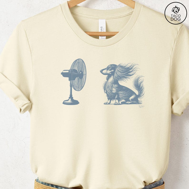 May include: Cream-colored t-shirt featuring a blue illustration of a dog with wind-blown fur next to a fan. The dog is a long-haired dachshund. The text "TACO DOG" is in a circle on the upper right.