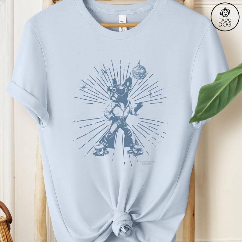 May include: Light blue t-shirt with a graphic of a dog wearing roller skates and holding a disco ball. The dog is wearing sunglasses and a disco outfit. The graphic is surrounded by a sunburst design.
