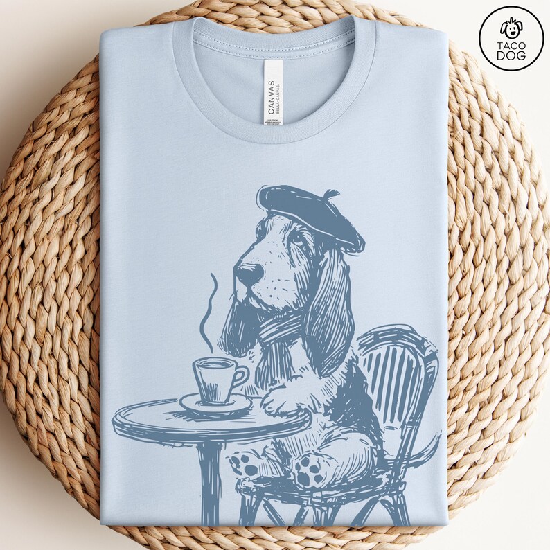 K&ouml;nnte beinhalten: Ein hellblaues T-Shirt mit einer Cartoon-Illustration eines Basset Hounds, der einen Barett tr&auml;gt und auf einem Stuhl mit einer Tasse Kaffee sitzt. Der Hund schaut nach links und tr&auml;gt einen Schal um den Hals. Der Stuhl ist gestreift und hat einen runden Tisch davor.