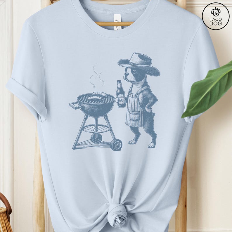 Peut inclure: Un t-shirt bleu clair avec un graphique d'un chien portant un chapeau de cowboy et un tablier grillant des saucisses. Le chien tient une bouteille de bi&egrave;re. Le texte "Taco Dog" est dans le coin sup&eacute;rieur droit de l'image.