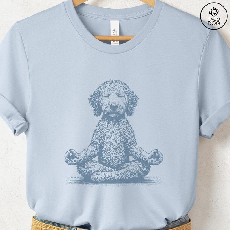 May include: Light blue t-shirt featuring a detailed illustration of a dog in a seated yoga pose. The dog is depicted with closed eyes and crossed legs, suggesting meditation. The design is in a monochromatic style, with the dog's fur rendered in intricate detail.