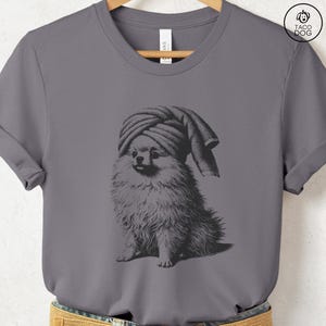 May include: Gray t-shirt featuring a black and white illustration of a Pomeranian dog wearing a towel on its head. The dog is sitting, with its tongue slightly visible. The shirt has a crew neck and short sleeves.