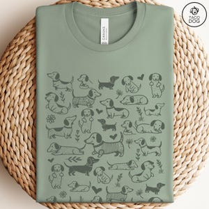 May include: A sage green t-shirt with a repeating pattern of black dachshund dogs in various poses. The shirt has a crew neck and short sleeves.
