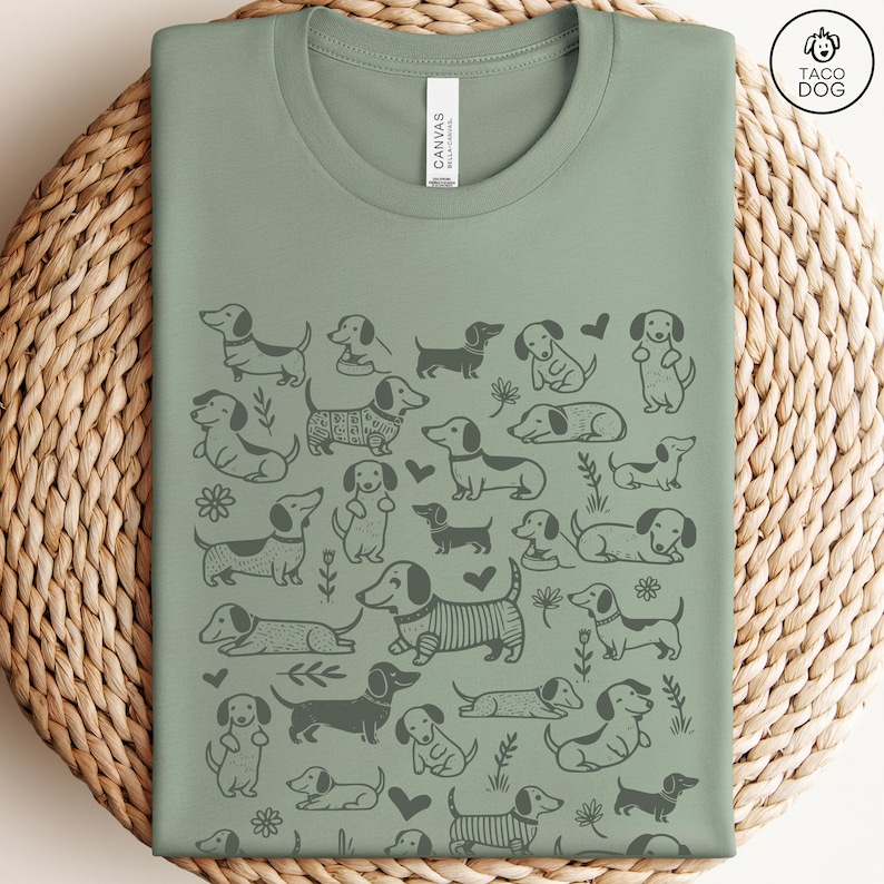 May include: A sage green t-shirt with a repeating pattern of black dachshund dogs in various poses. The shirt has a crew neck and short sleeves.