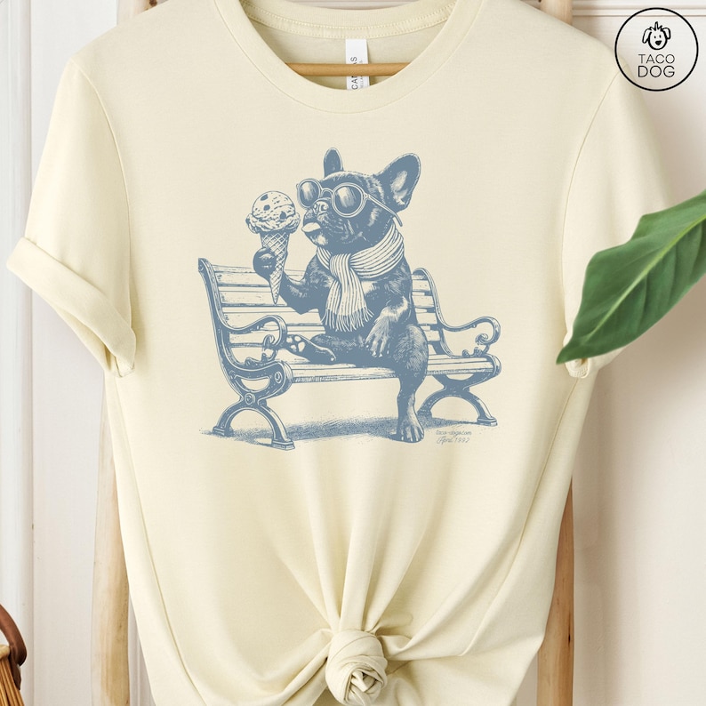 May include: Cream-colored t-shirt featuring a blue illustration of a French Bulldog wearing sunglasses and a scarf, holding an ice cream cone while sitting on a park bench. The shirt has a relaxed fit and a crew neck.