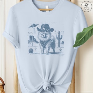 Puede incluir: Una camiseta azul claro con un gr&aacute;fico de un perro con un sombrero de vaquero y una bandana en un paisaje des&eacute;rtico. El perro es de raza Pomerania. El gr&aacute;fico es de estilo vintage con un aspecto desgastado. La camiseta es de estilo unisex.