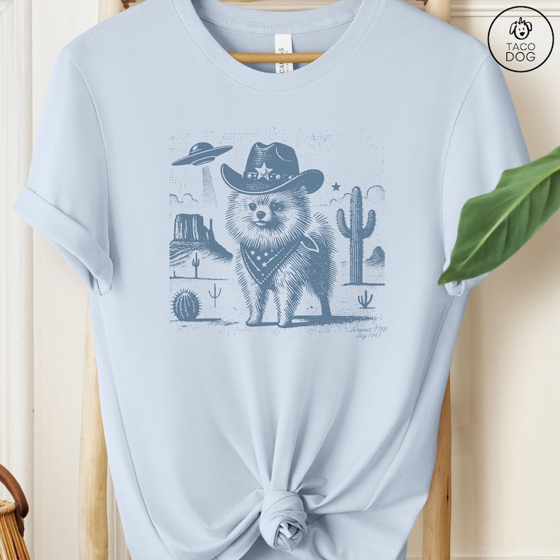 Puede incluir: Una camiseta azul claro con un gr&aacute;fico de un perro con un sombrero de vaquero y una bandana en un paisaje des&eacute;rtico. El perro es de raza Pomerania. El gr&aacute;fico es de estilo vintage con un aspecto desgastado. La camiseta es de estilo unisex.