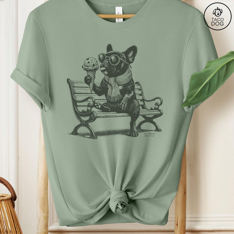 May include: Sage green t-shirt featuring a black and white illustration of a French Bulldog wearing sunglasses and a scarf, holding an ice cream cone while sitting on a park bench. The design includes the text "TACO DOG".