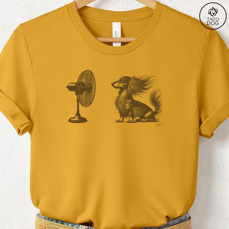 May include: Mustard yellow t-shirt featuring a detailed illustration of a long-haired dachshund enjoying the breeze from a fan. The dog's fur is blowing in the wind. The shirt has a crew neck and short sleeves.