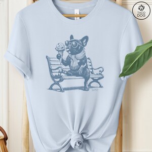 May include: Light blue t-shirt featuring a blue illustration of a French Bulldog wearing sunglasses and a scarf, holding an ice cream cone while sitting on a park bench. The shirt has a tied knot at the bottom.