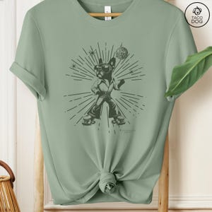 May include: A green t-shirt with a graphic of a dog wearing roller skates and holding a disco ball. The dog is wearing a 70s-style outfit and has a disco ball in its mouth. The graphic is surrounded by a sunburst design.