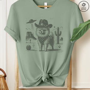 Puede incluir: Una camiseta verde con una ilustraci&oacute;n en blanco y negro de un perro con un sombrero de vaquero y una bandana en un paisaje des&eacute;rtico. El perro es de raza Pomerania. El texto "Taco Dog" est&aacute; en la esquina superior derecha de la imagen.