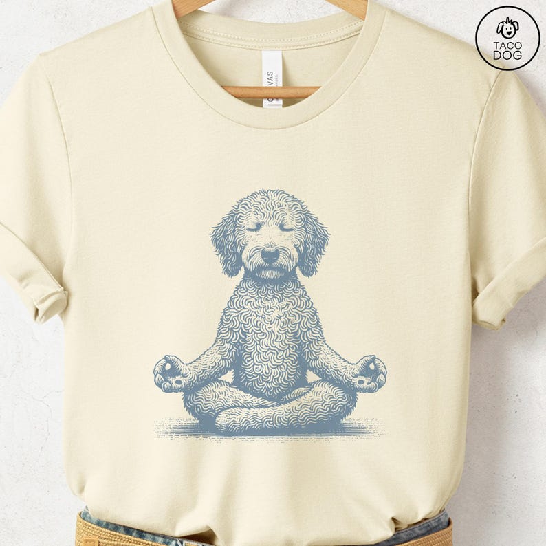 May include: Cream-colored t-shirt featuring a light blue illustration of a dog in a yoga pose. The dog is depicted with its eyes closed, sitting cross-legged in a meditative position. The shirt has short sleeves and a crew neck, with the words "TACO DOG" printed on the upper right.