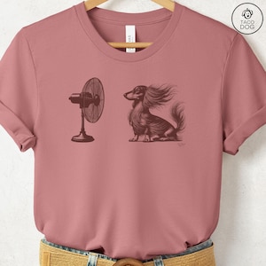May include: Dusty rose t-shirt featuring a detailed illustration of a long-haired dachshund with wind-blown fur, facing a vintage-style fan. The design is in sepia tones, creating a classic, artistic look. The shirt is a crew neck.