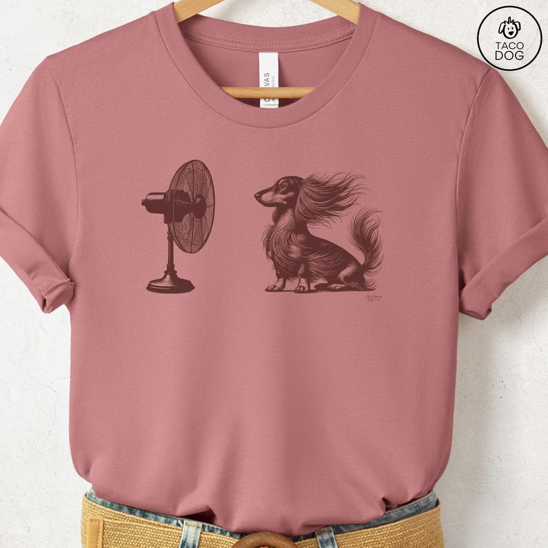 May include: Dusty rose t-shirt featuring a detailed illustration of a long-haired dachshund with wind-blown fur, facing a vintage-style fan. The design is in sepia tones, creating a classic, artistic look. The shirt is a crew neck.
