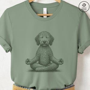 May include: Sage green t-shirt featuring a detailed illustration of a dog in a seated yoga pose. The dog has closed eyes and is in the lotus position. The shirt has a crew neck and short sleeves. The brand logo "TACO DOG" is in the upper right corner.