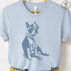 Puede incluir: Camiseta azul claro con un dise&ntilde;o de un Boston Terrier con gafas de esqu&iacute; y una bufanda. El perro est&aacute; representado sentado, en un tono azul monocrom&aacute;tico. La camiseta tiene cuello redondo y mangas cortas.