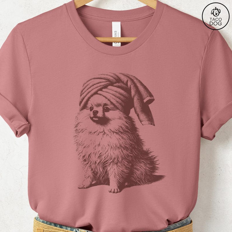 May include: Dusty rose t-shirt featuring a sepia-toned illustration of a Pomeranian dog wearing a towel on its head. The dog is sitting, with its fluffy fur detailed. The shirt has a crew neck and short sleeves. The Taco Dog logo is in the upper right corner.