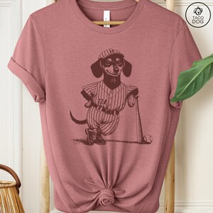 May include: A pink t-shirt with a graphic of a dachshund wearing a baseball uniform and holding a bat. The dachshund is wearing a baseball cap with the letter "H" on it. The text "124" is on the uniform.