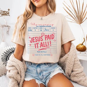 May include: A beige t-shirt with a pink and red graphic that says "Why we paying all these bills when Jesus paid it all!" The graphic is styled like a receipt with the words "Paid in full!" and "Thank you. Tell your friends." at the bottom.