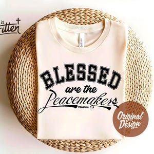 Puede incluir: Camiseta color crema con la frase "BLESSED are the Peacemakers" en negro. La palabra "BLESSED" está en una fuente universitaria. La camiseta está sobre una superficie circular y tejida. También se ve el texto "Matthew 5:9".