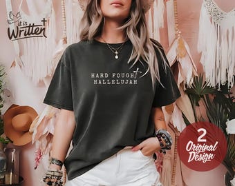 May include: Dark gray t-shirt with the text "HARD FOUGHT HALLELUJAH" and a cross graphic. The shirt is a relaxed fit and has a vintage look. A red circle with "2 Original Design" is on the bottom right.