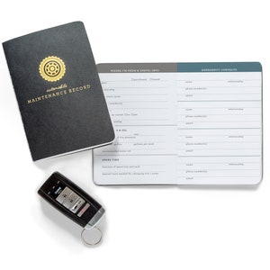 Puede incluir: Un libro de registro de mantenimiento de automóviles negro y gris con un logotipo de rueda dorado. El libro está abierto en páginas con secciones para contactos de emergencia, de dónde es el automóvil y información útil. Una llave de coche negra está en primer plano.