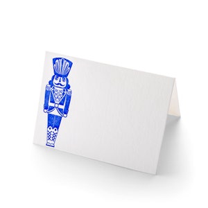 Op de afbeelding: Een witte placecard met een blauwe illustratie van een notenkraker. De notenkraker draagt een blauwe hoed, een blauw jasje en een blauwe broek. De notenkraker heeft een witte snor en een witte baard.