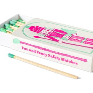 May include: A white matchbox with a pink and green illustration of a toy soldier and the text "Fun and Fancy Safety Matches." The matches have green tips. One match is separate.
