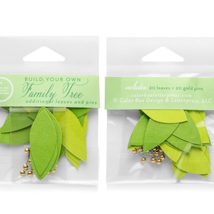 May include: Two clear plastic bags containing green paper leaves and gold pins. The bag on the left has a label that reads "BUILD YOUR OWN Family Tree additional leaves and pins". The bag on the right has a label that reads "includes 20 leaves + 20 gold pins colorboxletterpress.com © Color Box Design & Letterpress, LLC".