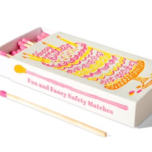 Puede incluir: Una caja de cerillas blanca con un diseño de tarta de cumpleaños colorido y el texto "Happy Birthday" y "Fun and Fancy Safety Matches". La caja contiene cerillas con punta rosa, con una cerilla fuera.