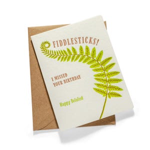 May include: A cream-colored greeting card with a brown kraft envelope. The card features a green fern illustration and brown text that reads FIDDLESTICKS! I MISSED YOUR BIRTHDAY, with the green text Happy Belated printed at the bottom.