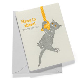 May include: A greeting card with a light cream background featuring a gray cat hanging from a yellow rope. The text reads "Hang in there! You've got this." in yellow and gray font. The card is positioned on top of a plain white envelope.