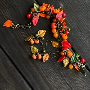 May include: A fall-themed bracelet and earrings set featuring miniature pumpkins, leaves, and beads in shades of orange, red, green, and gold. The bracelet is made with a chain and the earrings are dangle style.