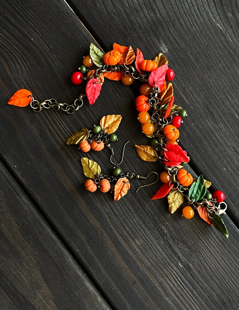 May include: A fall-themed bracelet and earrings set featuring miniature pumpkins, leaves, and beads in shades of orange, red, green, and gold. The bracelet is made with a chain and the earrings are dangle style.