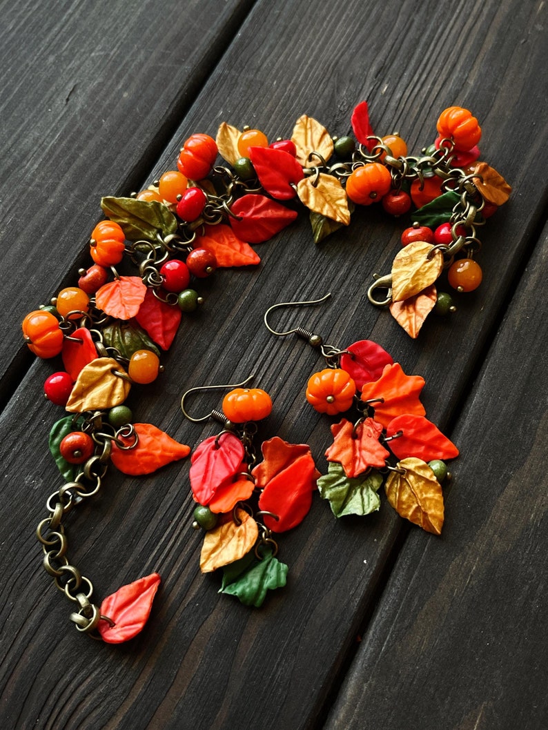 May include: A set of fall-themed jewelry featuring a bracelet and earrings. The bracelet and earrings are made with polymer clay and feature small pumpkins, leaves, and beads in shades of orange, red, green, and gold.