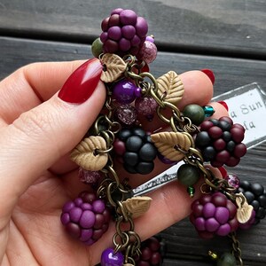 May include: A handcrafted charm bracelet featuring polymer clay blackberries in shades of purple and black. The bracelet includes small beads, leaf accents, and is connected by antique bronze chains. The design is a nature-inspired accessory.