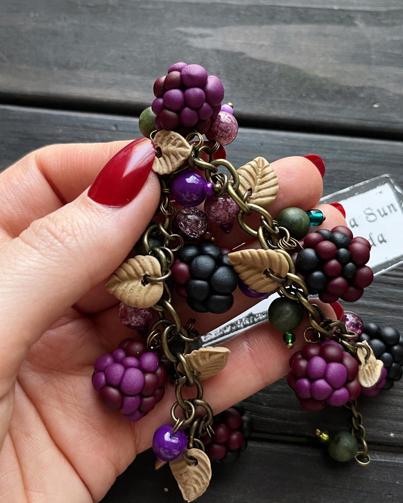 May include: A handcrafted charm bracelet featuring polymer clay blackberries in shades of purple and black. The bracelet includes small beads, leaf accents, and is connected by antique bronze chains. The design is a nature-inspired accessory.
