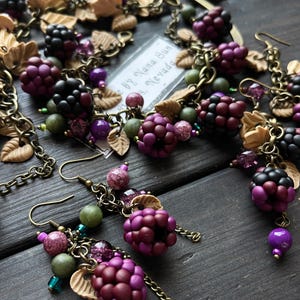 May include: A handcrafted jewellery set with a blackberry design, including a necklace and earrings. The set features dark purple and black berry beads, tan leaf details, and green and purple beads. The pieces are linked with bronze-toned chains and hooks.