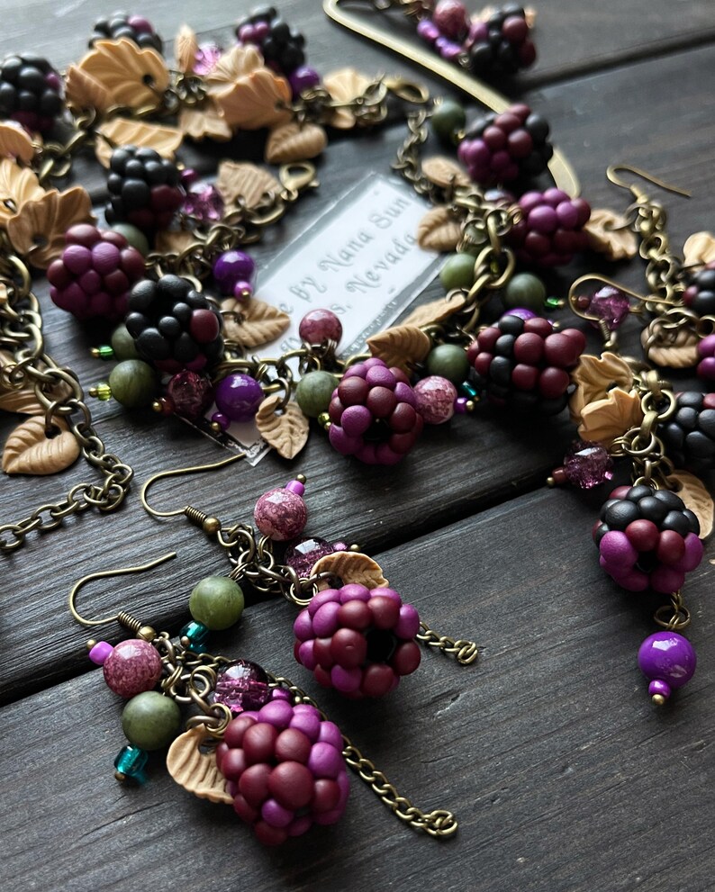 May include: A handcrafted jewellery set with a blackberry design, including a necklace and earrings. The set features dark purple and black berry beads, tan leaf details, and green and purple beads. The pieces are linked with bronze-toned chains and hooks.