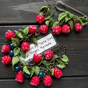 May include: Handmade jewelry set featuring red raspberry charms with green leaves and blue beads. The set includes earrings, a bracelet, and a brooch. The jewelry is made by Nana Sun, Las Vegas, Nevada.