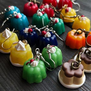 May include: A collection of colorful, miniature dessert-shaped earrings. The earrings are in various colors, including red, blue, green, yellow, and orange, with white frosting and a red cherry on top. Some earrings have a chocolate-colored top.